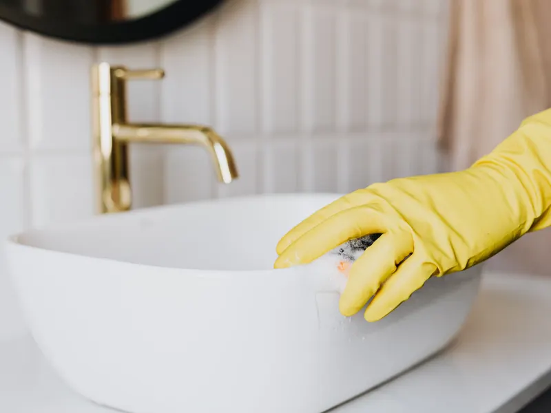 A gloved hand carefully wiping a white bathroom basin beside a brass faucet