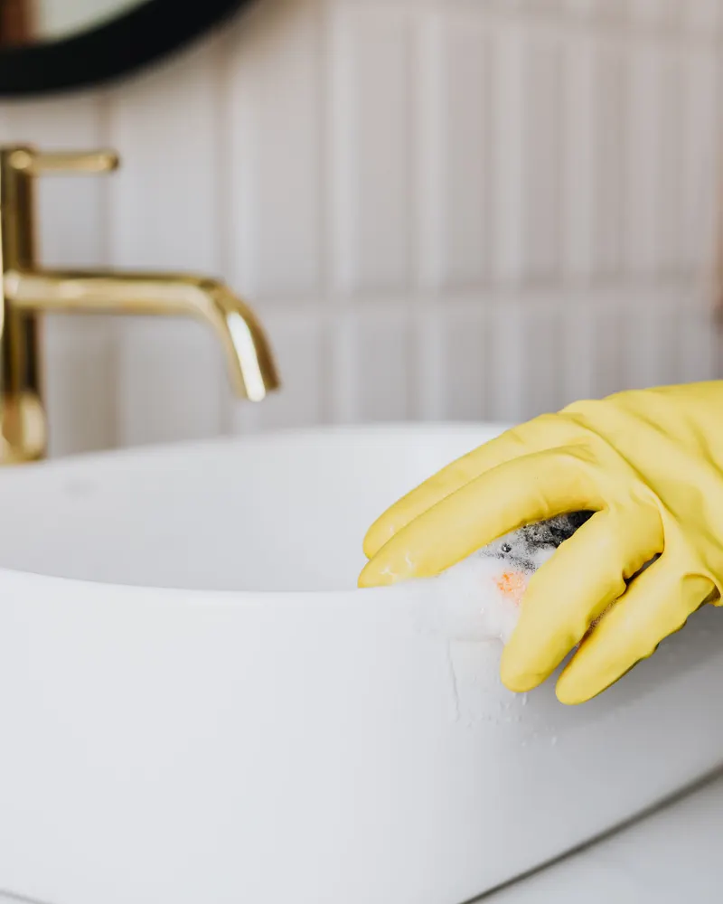 A gloved hand carefully wiping a white bathroom basin beside a brass faucet